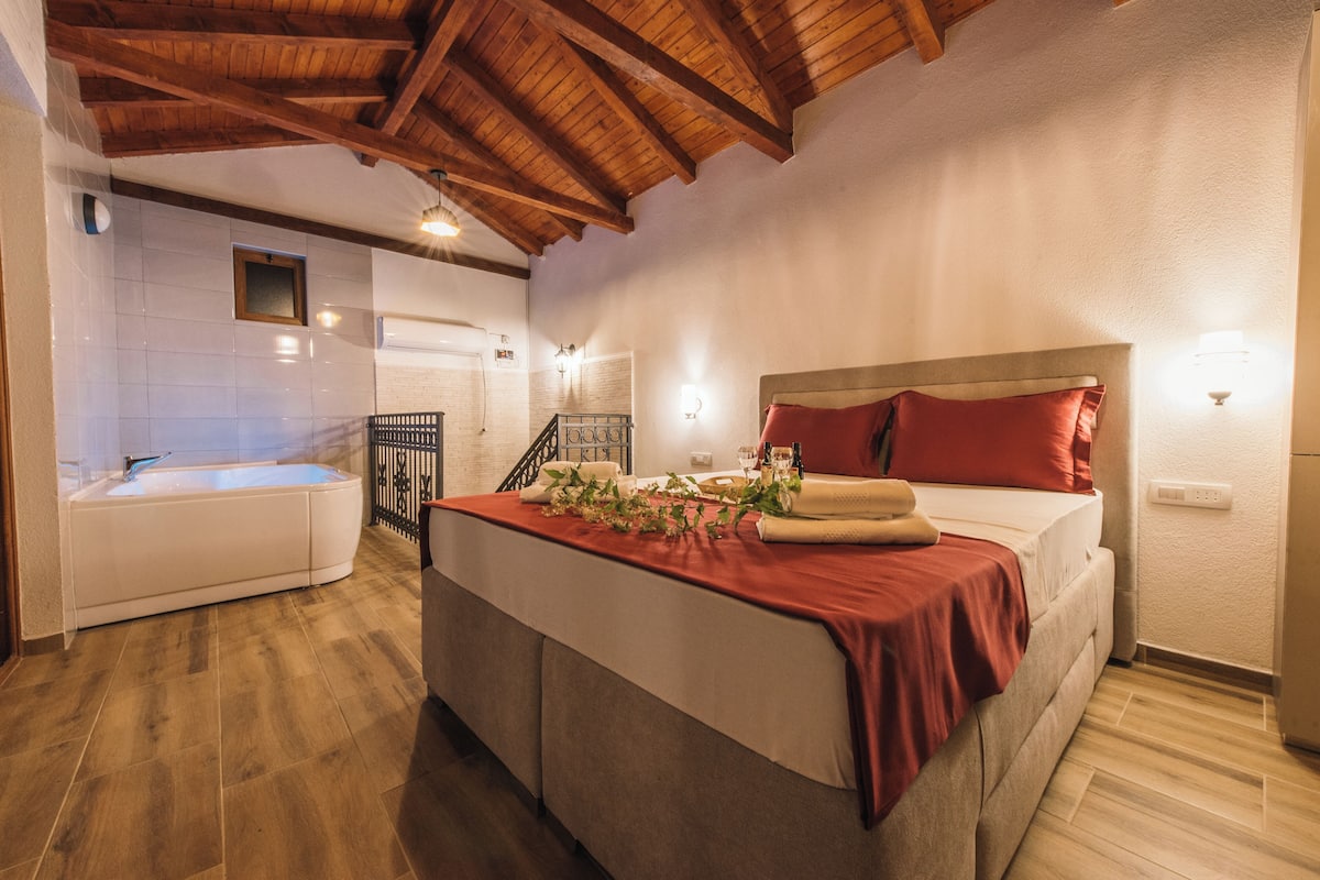 Ein Schlafzimmer mit Bett, roter Decke, weißem Kopfende, Holzboden und einem Badezimmer mit Badewanne.