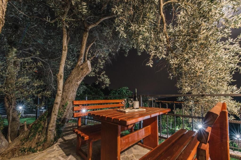 An outdoor seating area with a wooden table and benches, surrounded by trees and a metal fence.