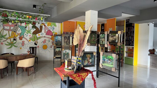 Een kamer met betegelde vloer, een zithoek met stoelen, een tafel met een rode tafelkleed, en een muur met kleurrijke kunstwerken en een hangende plant.
