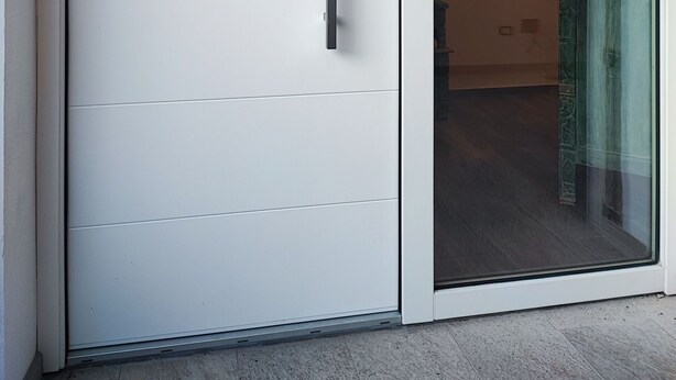 Una puerta corredera blanca con manija y cerradura, que da a un espacio con puerta de vidrio y planta decorativa.