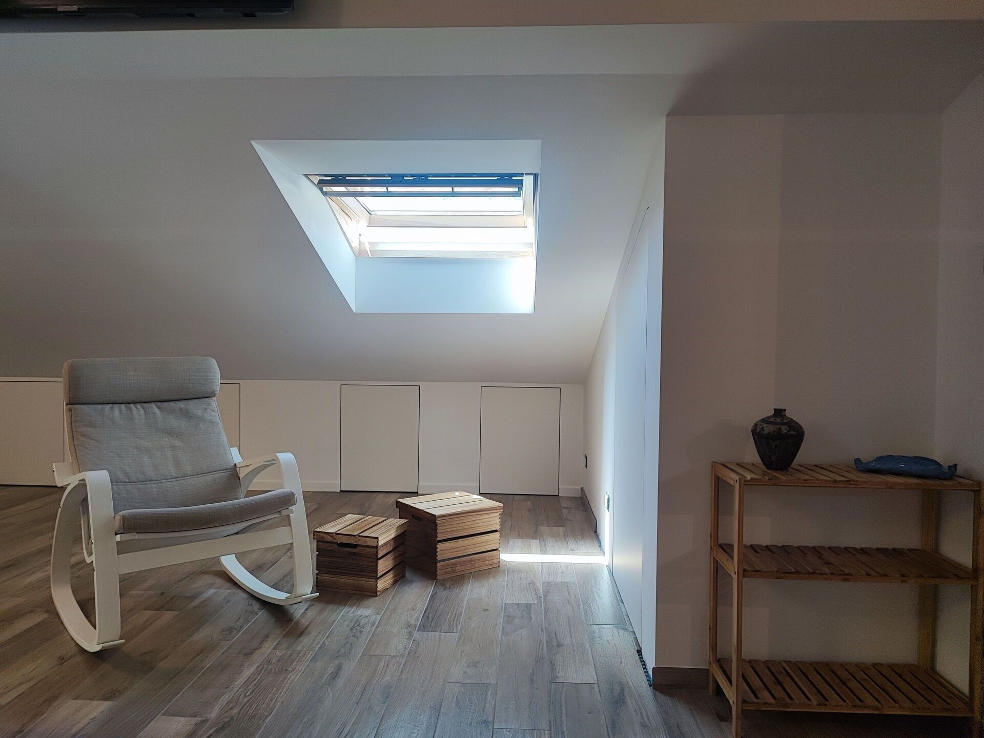 A room with a skylight, a rocking chair, wooden crates, and a shelf with a vase.