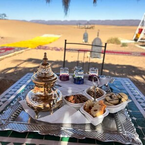 Une table sur laquelle sont disposés un plateau en argent, un récipient décoratif en argent et de petits pots en verre contenant des éléments aux couleurs vives, le tout sur fond de paysage désertique.