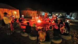 Eine Gruppe von Menschen sitzt nachts am Lagerfeuer, im Hintergrund sind Zelte zu sehen.