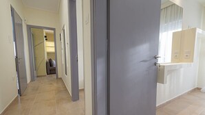 A hallway with a grey door, a bathroom with a mirror, and a tiled floor. - Happy Ellen Seafront 1 by Travel Pro Services (Nea Plagia)