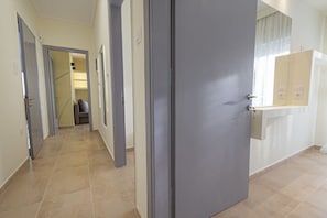 A hallway with a grey door, a bathroom with a mirror, and a tiled floor. - Happy Ellen Seafront 1 by Travel Pro Services (Nea Plagia)