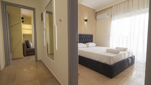 A bedroom with a bed, a large mirror, a door leading to a hallway, and a window with curtains. - Happy Ellen Seafront 1 by Travel Pro Services (Nea Plagia)