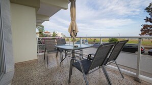 A balcony with a table and chairs, an umbrella, and a view of the street and beach. - Happy Ellen Seafront 1 by Travel Pro Services (Nea Plagia)
