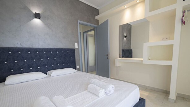 A modern hotel room with a large bed, a built-in bench, a wall-mounted mirror, and a small bathroom area. - Happy Ellen Seafront 4 by Travel Pro Services (Nea Plagia)