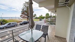 A balcony with a glass table and chairs, overlooking a street with cars and a beach. - Happy Ellen Seafront 4 by Travel Pro Services (Nea Plagia)
