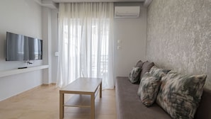Un soggiorno moderno con una TV a schermo piatto, un tavolo di legno, un divano con cuscini decorati e un'ampia finestra con tende trasparenti.