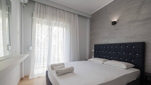 Une chambre d’hôtel moderne avec un grand lit, du linge de lit blanc, une tête de lit sombre et des rideaux transparents.