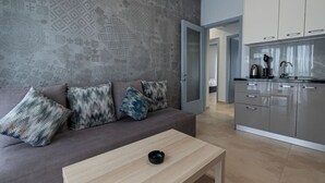 Un salon moderne avec un canapé gris, des coussins à motifs, une table basse en bois et une cuisine aux meubles gris.