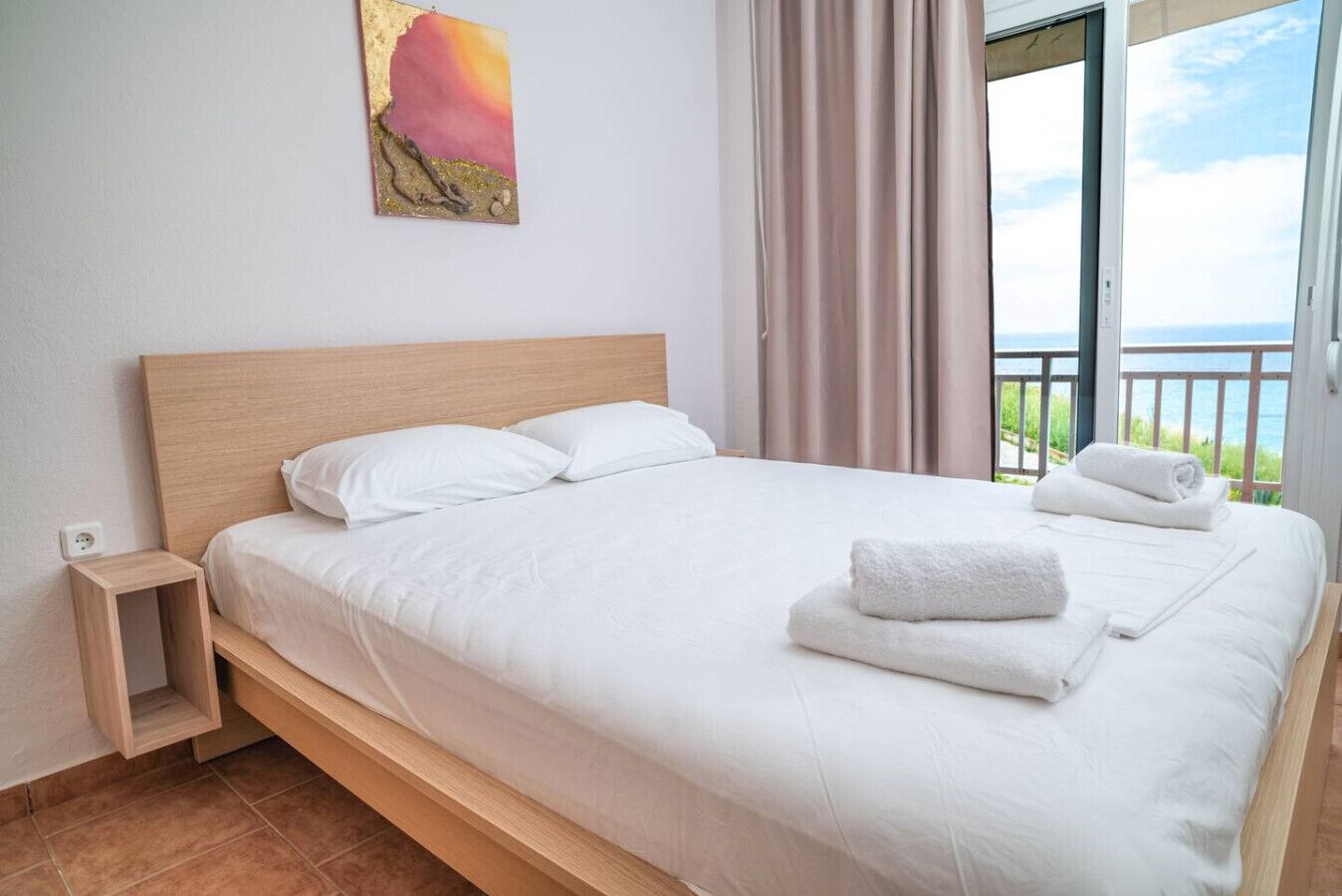 A hotel room with a bed, a small wooden nightstand, a painting on the wall, and a view of the sea through the window.