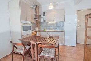 A compact kitchen with a dining table and chairs, a built-in oven, a refrigerator, and a hanging light fixture. - Seagull House by RentalsPro - Nea Potidaia (Nea Potidaia)