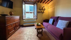 A cozy room with a wooden dresser, a purple sofa, a wooden table, and a window with a view of a building. - Lovely apartment for 3 people with WIFI, A/C, terrace, pets allowed and panoramic view (Cortona)