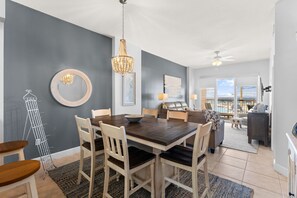 A dining area with a wooden table and chairs, a chandelier, and a view of the balcony. - NEW Sunrise Beach 510! FREE Beach Service, Heated Pool, FREE Golf & Perks! (Panama City Beach)