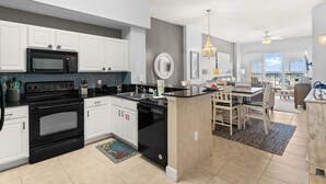 A modern kitchen with white cabinets, a black stove, and a dishwasher. There is a dining area with a table and chairs. - NEW Sunrise Beach 510! FREE Beach Service, Heated Pool, FREE Golf & Perks! (Panama City Beach)