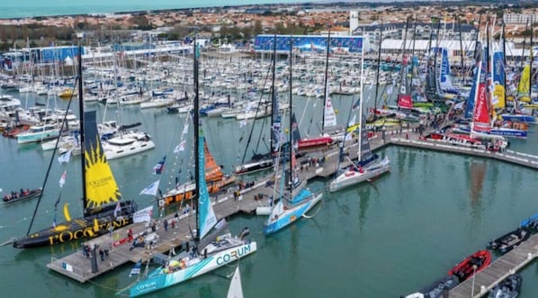 Een jachthaven met diverse boten afgemeerd, een steiger met mensen erop en een stadsgezicht op de achtergrond.