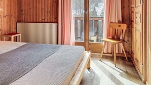 Et trægulvs værelse med seng, stol, radiator og et vindue med udsigt til træer.
