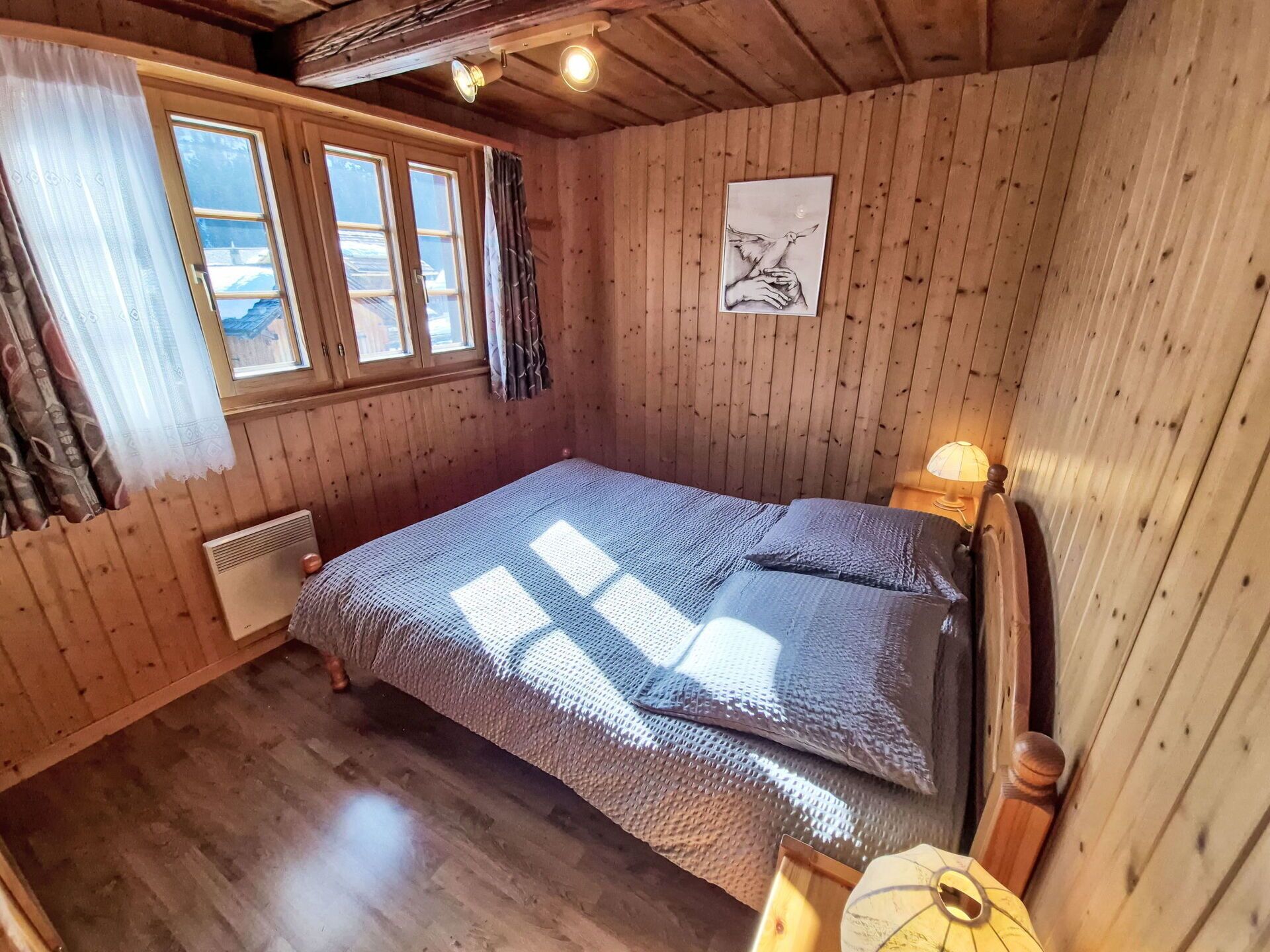 A wooden-paneled room with a bed, a window with curtains, a radiator, and a wall-mounted lamp.