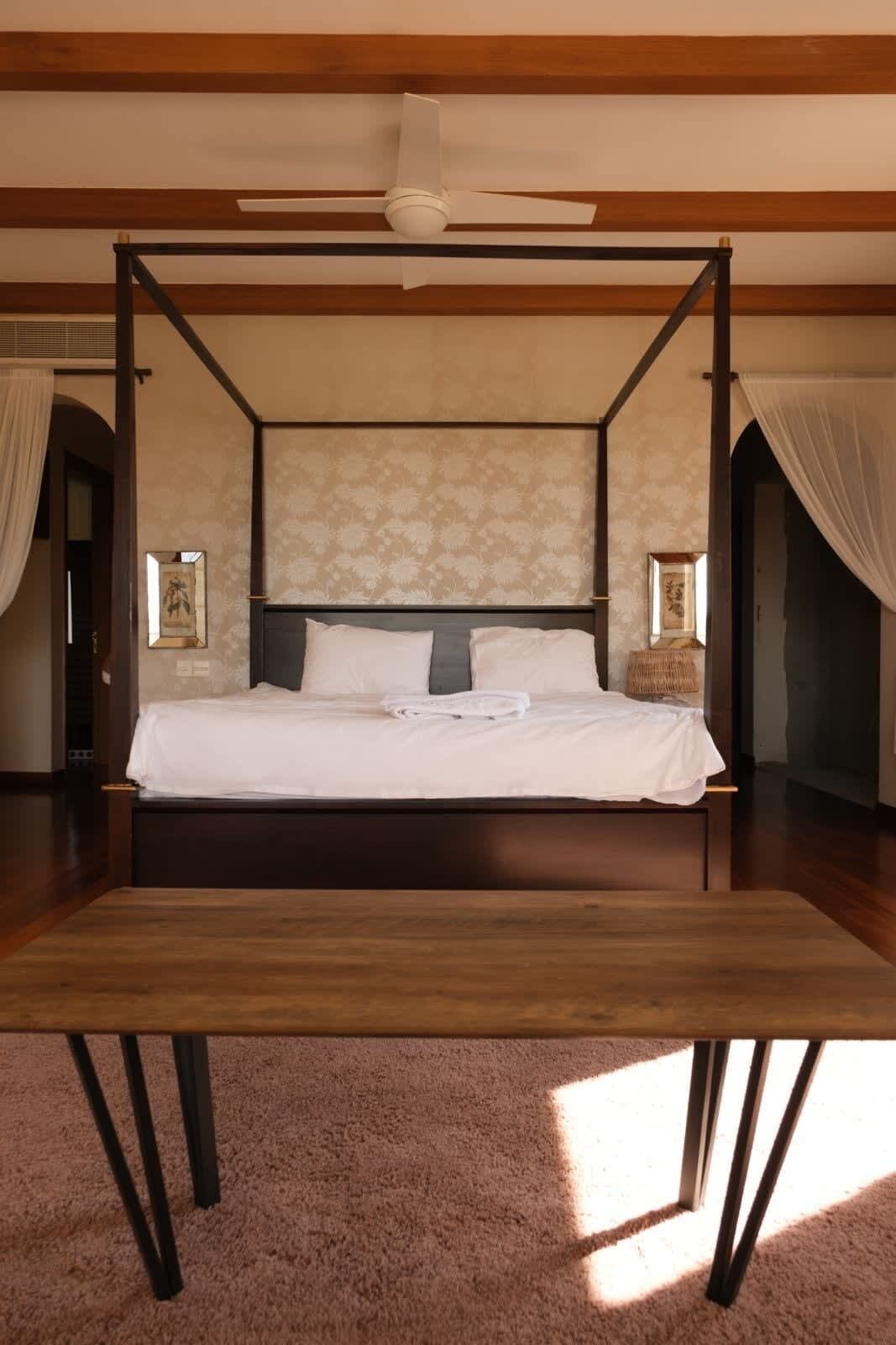 Una cama con dosel y estructura de madera, cabecera con estampado floral y un banco frente a la cama.