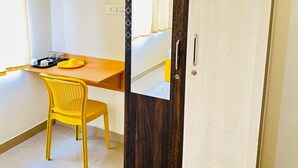 A room with a wardrobe, a mirror, a yellow chair, a wooden table, and a small shelf with items. - SM Stay Inn (Bengaluru)