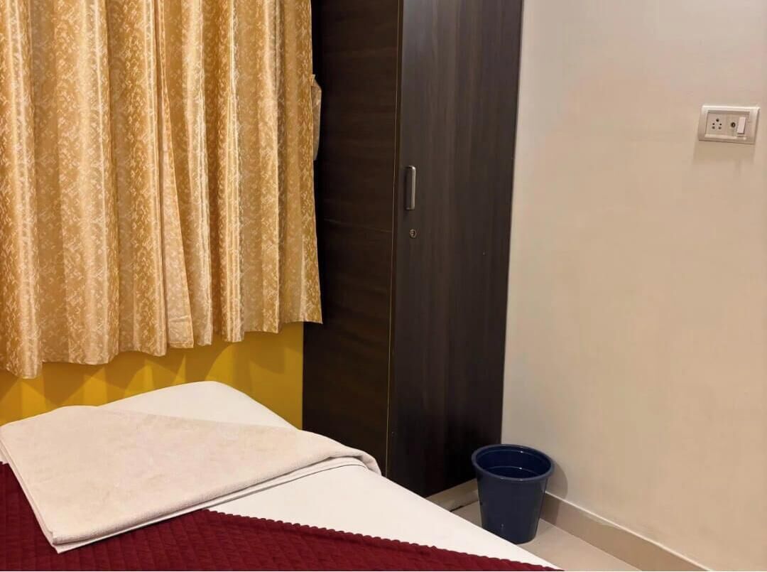A room with a bed, a curtain, a wardrobe, a blue bucket, and a wall socket.