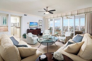 A spacious living room with a large glass coffee table, comfortable seating, and a city view through large windows. - Regatta - Walk to the Beach! (Naples)