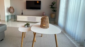 Una sala de estar moderna con un televisor de pantalla plana, dos mesas auxiliares con adornos, un sofá blanco y un ventanal con cortinas de encaje.
