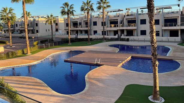 Zona de piscina con varias piscinas, palmeras y edificios modernos al fondo.