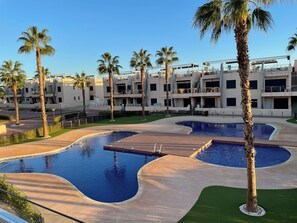 Zona de piscina con varias piscinas, palmeras y edificios modernos al fondo.
