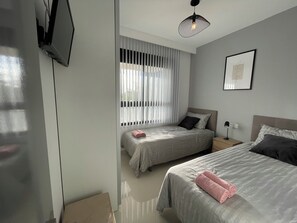 Ein modernes Schlafzimmer mit zwei Betten, einem an der Wand montierten Fernseher, einer Deckenleuchte und einem Fenster mit Jalousien.