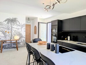 A modern kitchen with a black countertop, a dining area with a white table, and a mural of palm trees on the wall. - Villa Néréa - 4 Bedrooms and Pool - Near the Lagoon (Saint-Paul)