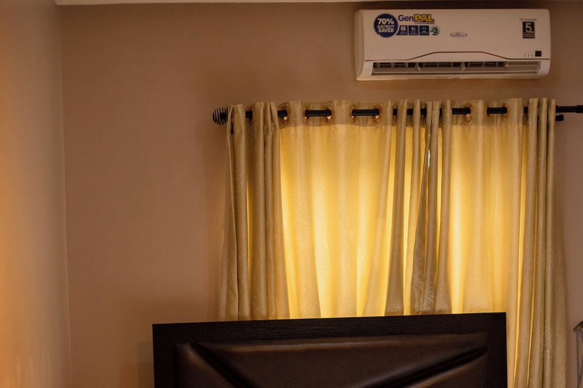 A hotel room with a bed, a headboard, a curtain, an air conditioning unit, and a chair.