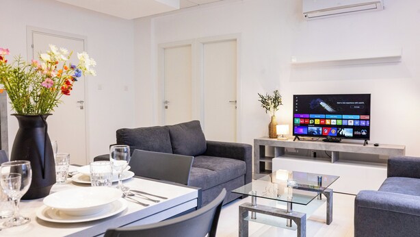 Ett modernt vardagsrum med en grå soffa, ett soffbord i glas, en matplats med ett vitt bord och stolar, samt en platt-TV som är monterad på väggen.