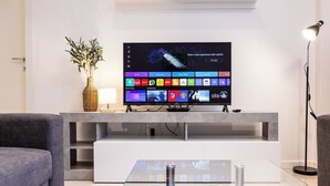 Ett modernt vardagsrum med en platt-TV, en vas med gröna växter, en lampa och ett soffbord i glas.