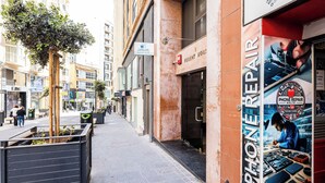 En stadsgata med en reparationsverkstad som heter ”PHONER REPAIR” på höger sida, en skylt med texten ”CAMILERI” ovanför, och ett träd i en blomlåda på vänster sida.