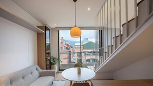 Uma sala de estar moderna com sofá, mesa redonda, luminária pendente e vista para o horizonte urbano através de amplas janelas.