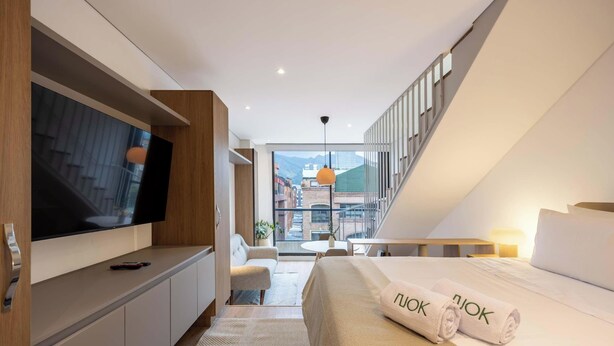 Quarto de hotel moderno com televisão, cama com toalhas, escrivaninha, cadeira e vista para edifícios através de grandes janelas.