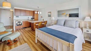 Habitaci ón de hotel compacta con kitchenette, una cama con colcha azul, zona de estar con una silla y un espejo grande sobre la cama.