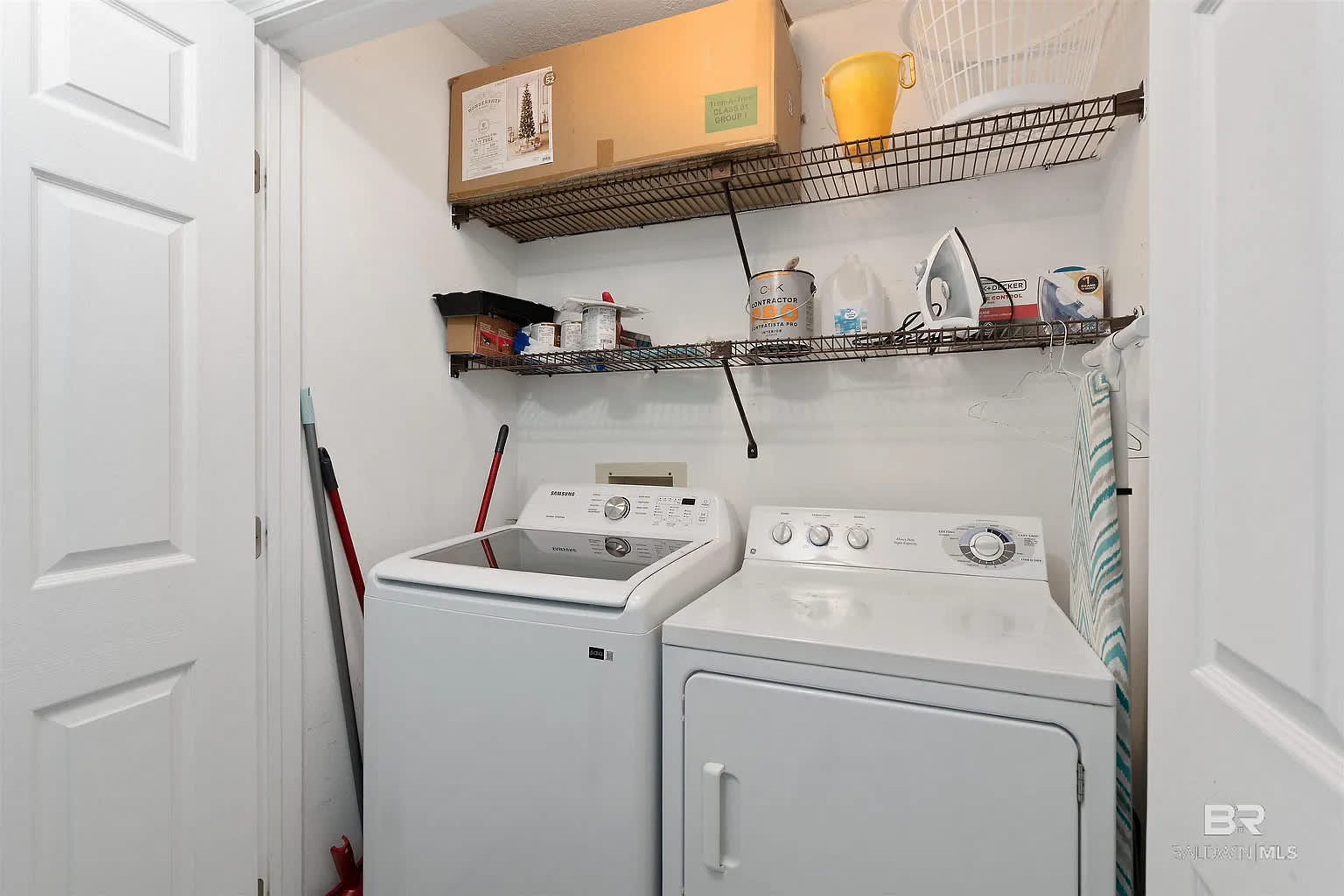 A laundry room with a front-loading washer and dryer, shelves above holding cleaning supplies, and a door leading to another room.