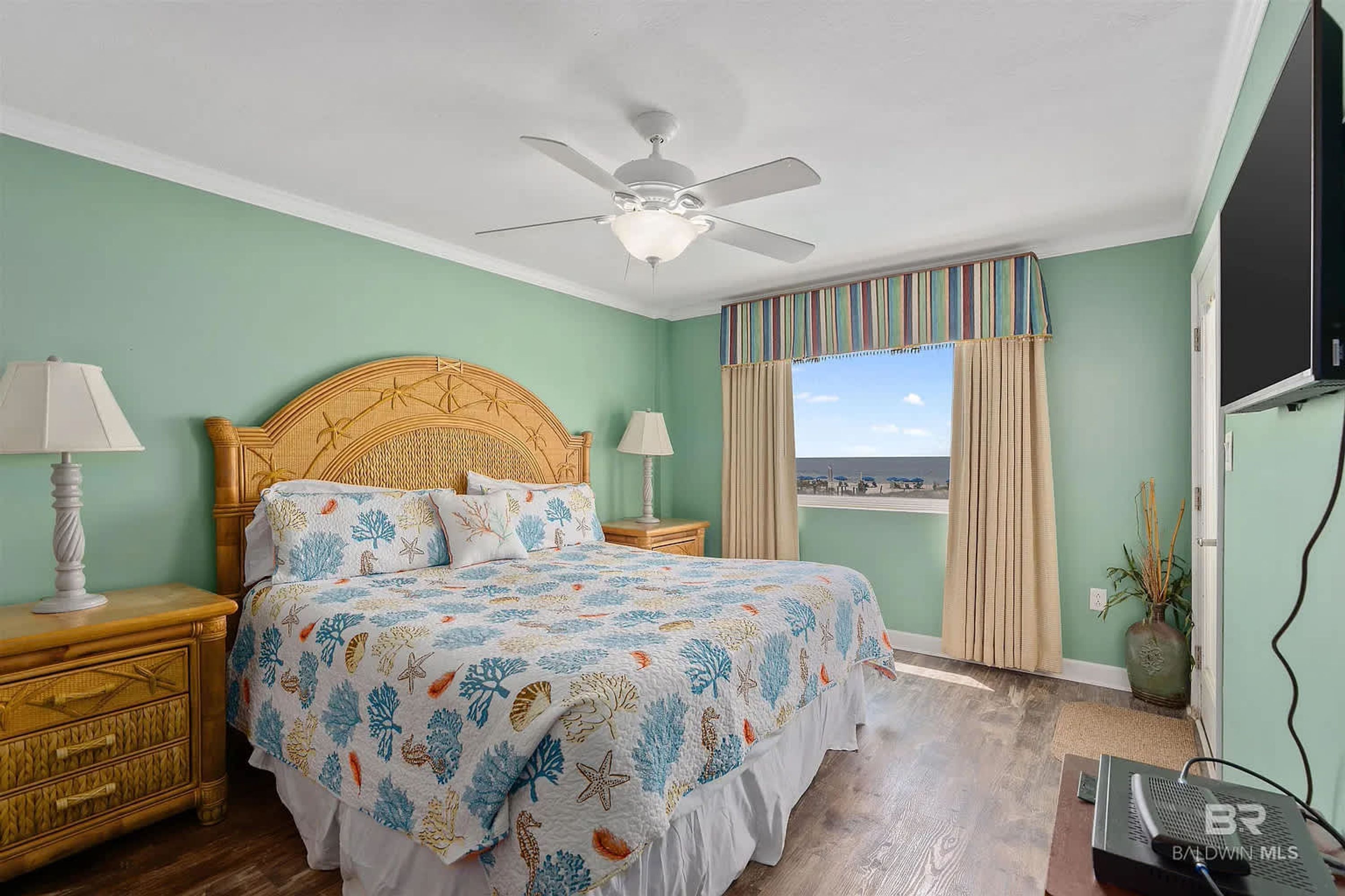Ein Schlafzimmer mit Bett, Deckenventilator, Fenster mit Vorhängen, Nachttisch und Lampe.
