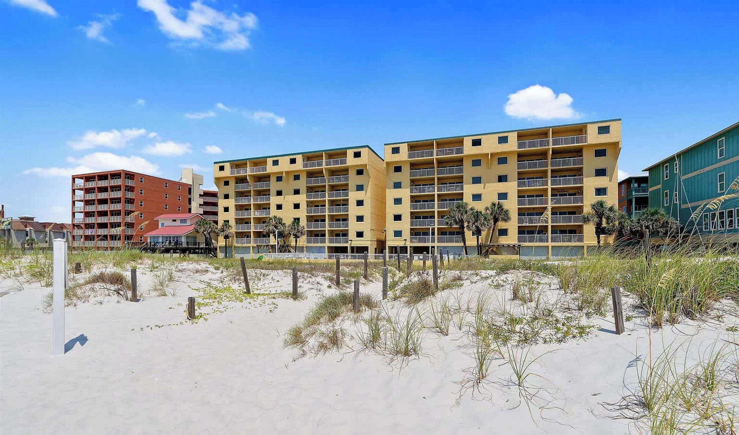 Un hôtel à plusieurs étages, doté de balcons, situé près d’une plage de sable où la végétation est clairsemée.