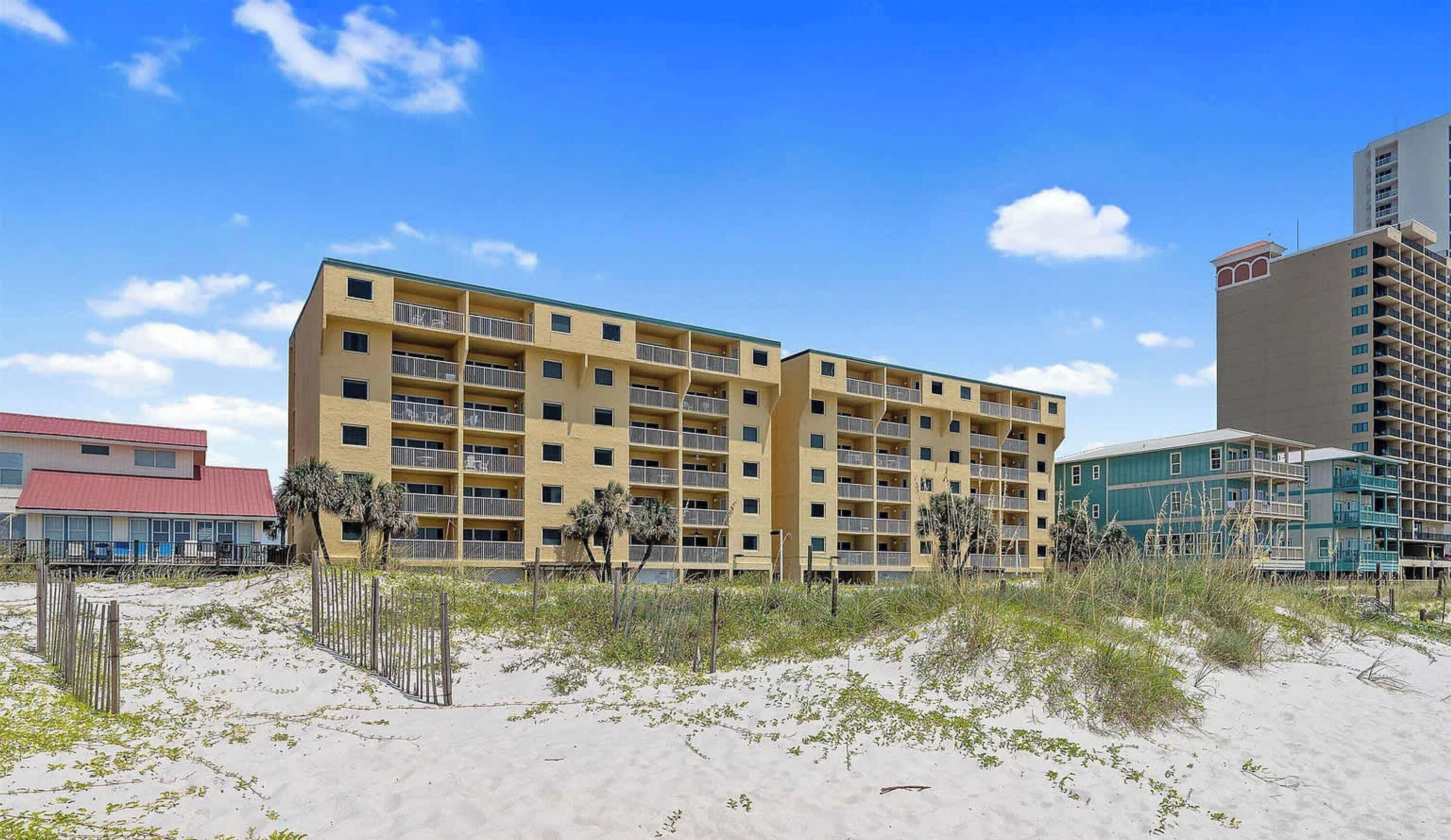 A multi-story hotel with balconies, located near a sandy beach with sparse vegetation.