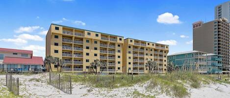 A multi-story hotel with balconies, located near a sandy beach with sparse vegetation.