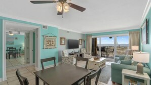 A living room with a TV, dining area, and balcony with ocean view. - The Big Surf II - Gulf View - Beach Pool and Fun (Gulf Shores)