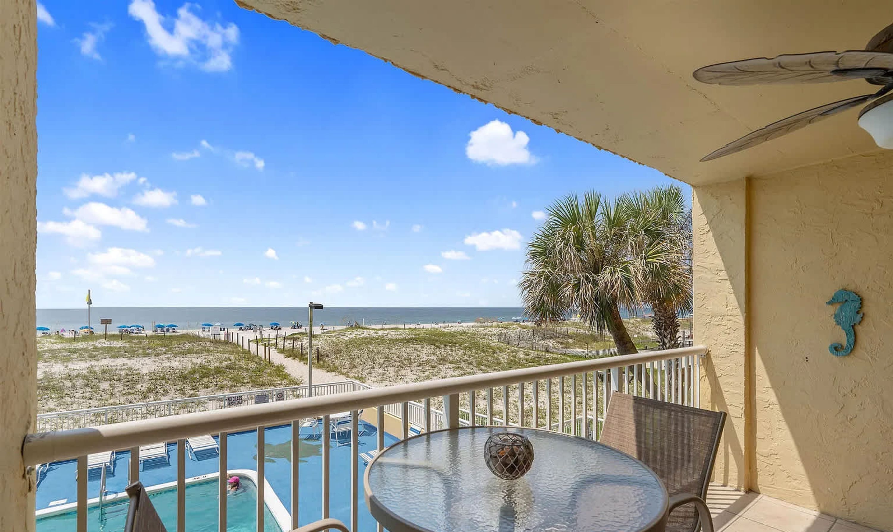 A balcony with a view of the beach, a pool, and a palm tree.