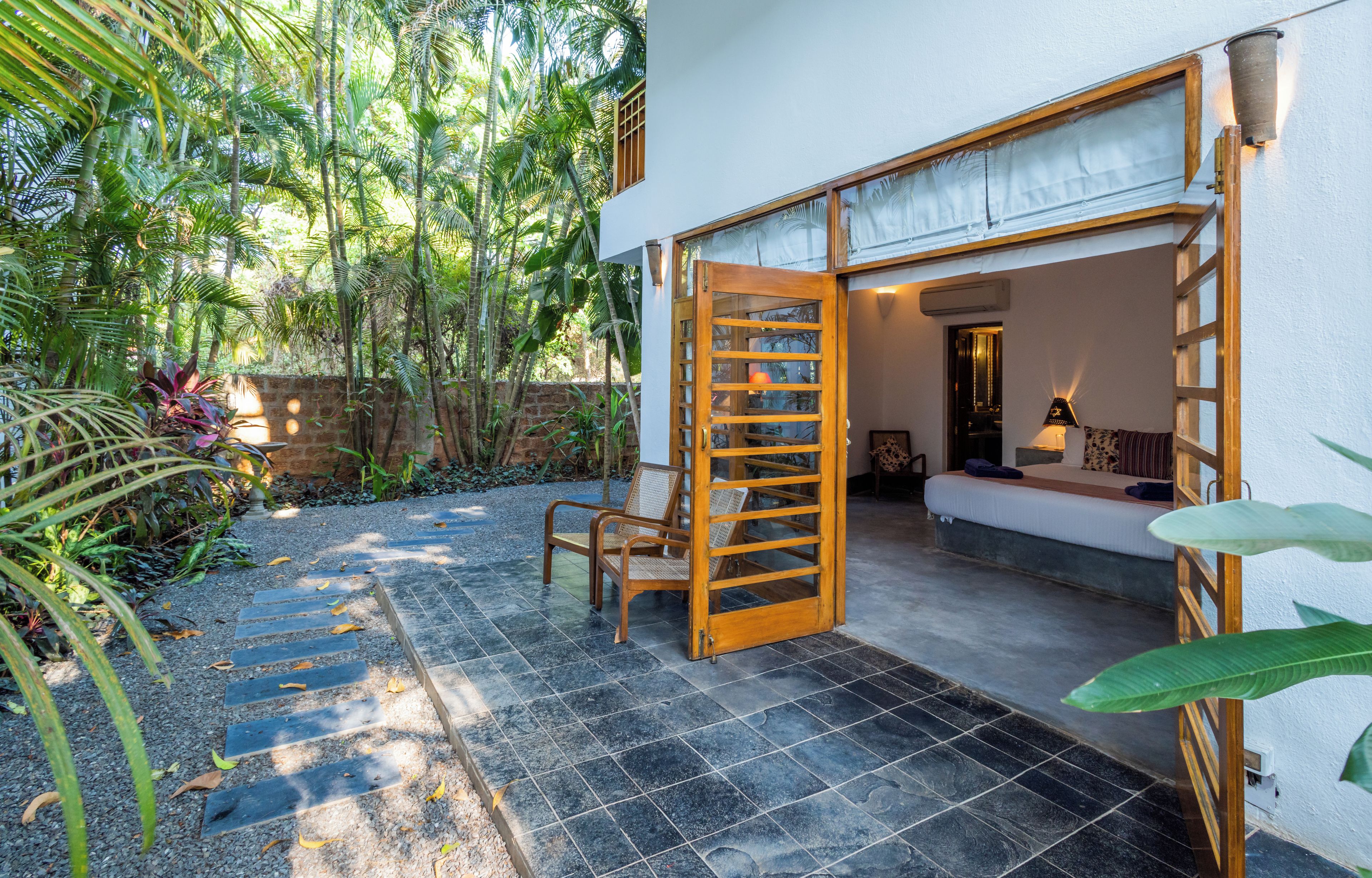 A room with a bed, a chair, and a wooden door leading to an outdoor area with a tiled floor and plants.