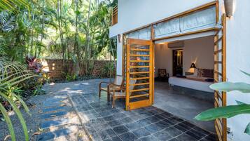 A room with a bed, a chair, and a wooden door leading to an outdoor area with a tiled floor and plants.