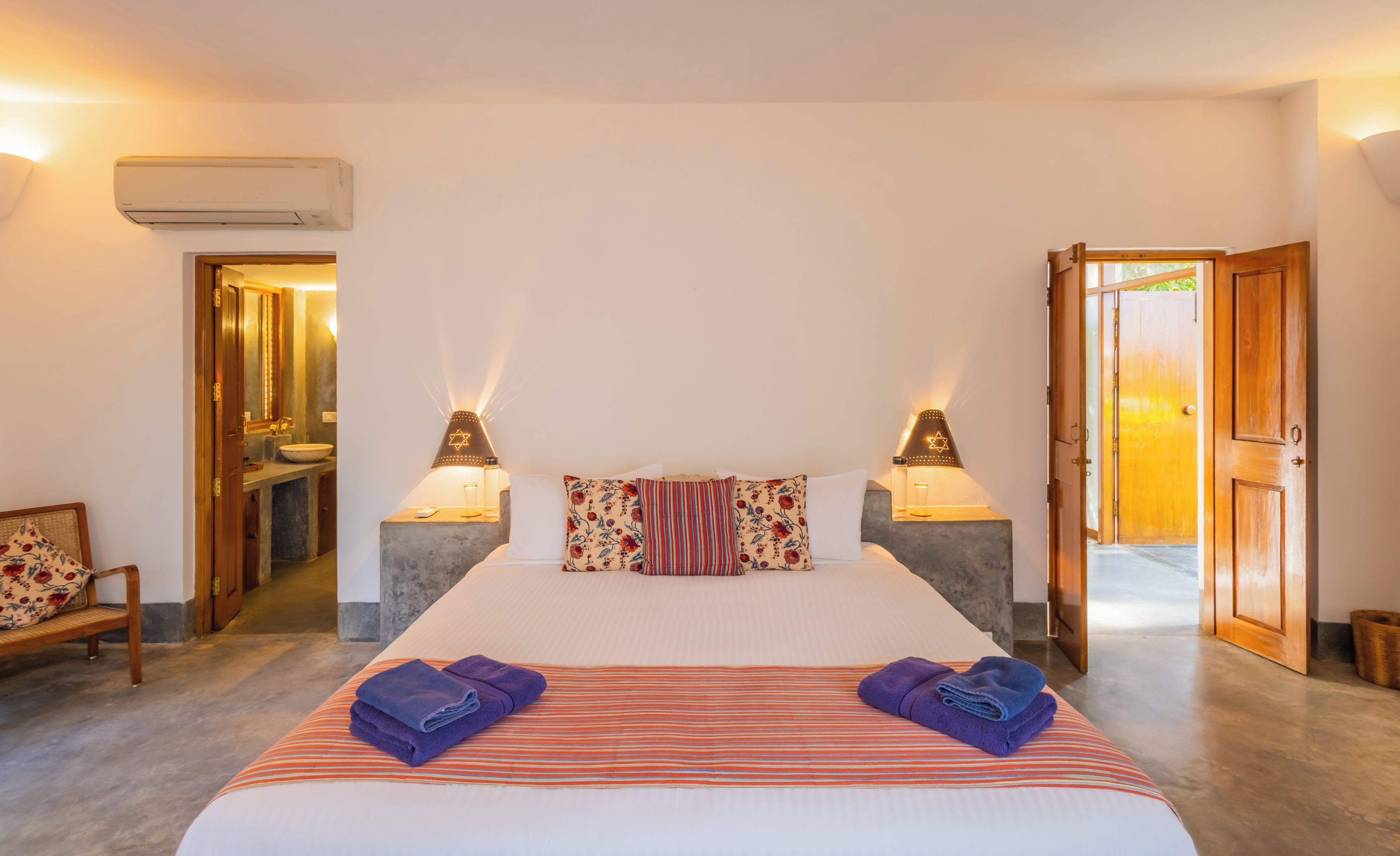Habitación de hotel con cama, dos lámparas de noche, baño visible a través de una puerta abierta y un banco de madera.